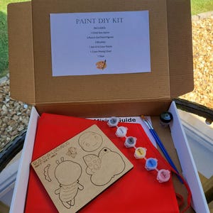 May include: A paint DIY kit in an open cardboard box. The kit includes a child-size apron, paint figures, brushes, paint, and a color mixing chart. The kit also includes a wooden cutout of a ladybug with the text "BUILD-A-LADYBUG PRINT IT - GLUE IT - HANG IT".