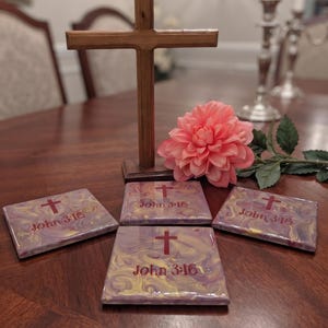 May include: A wooden cross stands behind four square coasters with a glossy, marbled purple and gold finish. Each coaster features a red cross icon and the text John 3:16 printed in red. A single pink dahlia flower rests near the base of the cross.