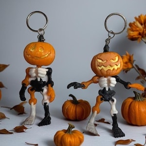 May include: Two Halloween-themed keychains featuring pumpkin heads and skeleton bodies. The pumpkins are orange with carved faces, and the skeletons are white, black, and orange. Small pumpkins and dried leaves are scattered around the keychains.