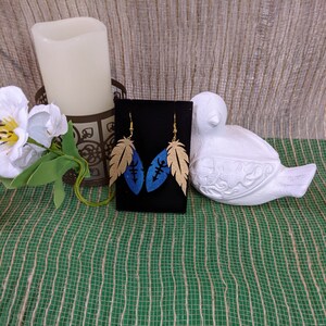 May include: A pair of gold and blue feather earrings with a geometric design. The earrings are on a black background.