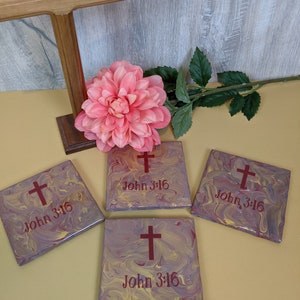 May include: Four square coasters with a marbled purple and yellow design. Each coaster features a red cross and the text "John 3:16".