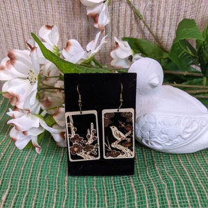 May include: A pair of wooden earrings with a black background and a white bird and floral design. The earrings are hanging from a black display card.