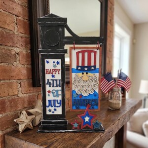 May include: A black wooden signpost with a hanging rectangular plaque featuring a stylized Uncle Sam figure, a top hat, and the word AMERICA. The post displays the text HAPPY 4TH OF JULY with blue and red stars. Includes three star-shaped accents.
