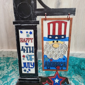 May include: A wooden sign with a black frame and a red, white, and blue Uncle Sam figure. The sign says "Happy 4th of July" and "America". The sign is decorated with stars and is attached to a black wooden post.