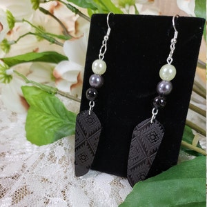 May include: A pair of dangle earrings with black and white geometric patterned pendants. The earrings feature a silver chain with three beads: a white pearl, a dark gray pearl, and a black bead.