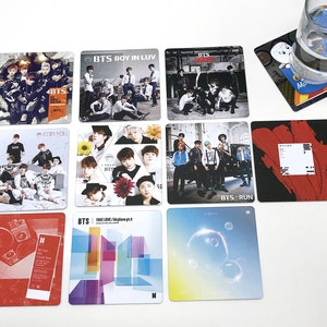 May include: A collection of square coasters featuring images of the K-pop group BTS. The coasters display album art and group photos, with text such as "BTS", "Boy in Luv", and "Run". A glass of water sits on a coaster with a cartoon character.