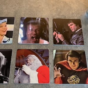 May include: Six square coasters with different images printed on them. The coasters feature images of a woman in a blue hat, a man in a helmet, a man holding a gun, a robot, a man in a Santa suit, and a man in a red cape.