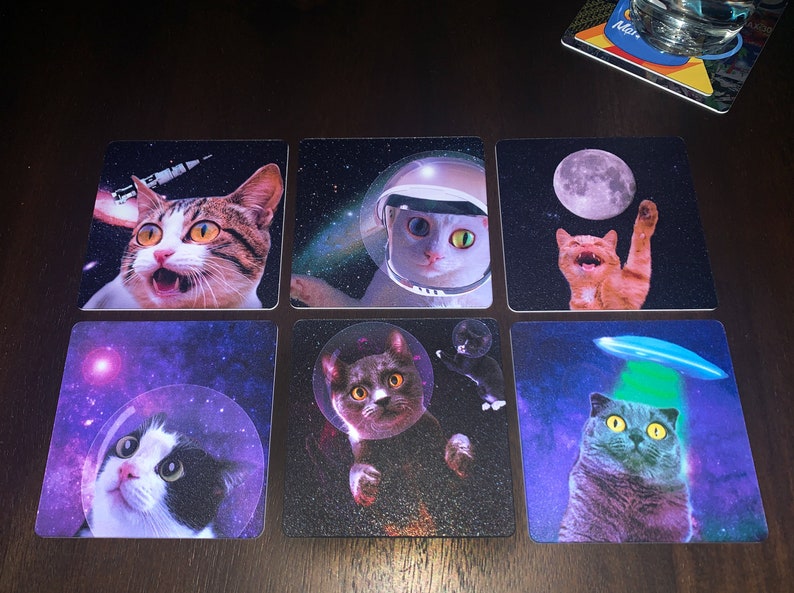 May include: Six square coasters with different images of cats in space. The coasters feature a cat in a spacesuit, a cat with a UFO over its head, a cat in a bubble, and a cat with a moon in the background.