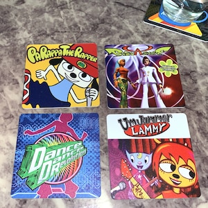 May include: Four colorful square coasters featuring characters from popular video games. The coasters feature Parappa the Rapper, Um Jammer Lammy, Dance Dance Revolution, and  'Bust a Groove'.