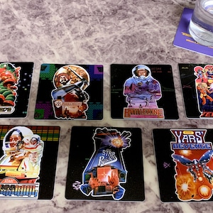 May include: A set of eight square coasters featuring retro video game graphics. The coasters are black with colorful images of classic arcade games, including Centipede, Battlezone, Tempest, Yar's Revenge, and Star Raider. The coasters are perfect for adding a touch of nostalgia to any home.