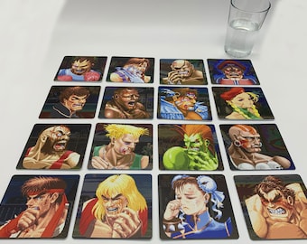 Könnte beinhalten: Ein Set mit 16 quadratischen Untersetzern mit Charakteren aus der Street Fighter Videospielserie. Jeder Untersetzer zeigt einen anderen Charakter mit einem pixeligen Design. Die Charaktere sind alle in verschiedenen Posen und Ausdrücken, einige mit Verletzungen. Die Untersetzer sind in einem Rastermuster auf einer weißen Oberfläche angeordnet.