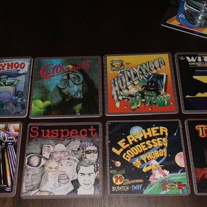 May include: Eight square coasters with different album covers. The coasters feature a variety of colors and designs, including a man in a diving suit, a cityscape, a bat, and a gate. The coasters are made of a hard material and have a smooth surface.