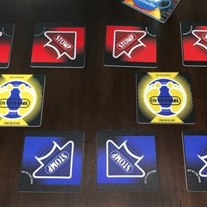May include: Eight square game tiles with different instructions. Four tiles are red with black arrows and the words "STOMP" on them. Two tiles are yellow with black footprints and the words "ON YOUR MARK" on them. Two tiles are blue with black arrows and the words "STOMP" on them.