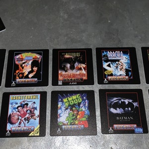 May include: Ten square black coasters with colorful images of classic video game cartridges. The coasters feature titles like Warbirds, Asteroids, Dracula, and Batman.
