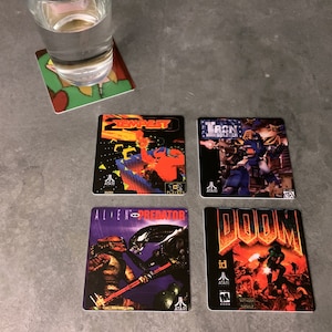 May include: Four square coasters featuring retro video game graphics. The coasters are black with colorful graphics. The coasters feature the games Tempest, Tron: Solar Sailer, Alien vs Predator, and Doom.