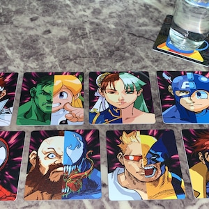 May include: Eight square coasters with a split image of two different comic book characters on each. The coasters have a black background with pink and purple streaks. The characters include: Iron Man and Ryu, Hulk and a girl with blonde hair, Chun-Li and a girl with green hair, Captain America and Mega Man, Spider-Man and Ryu, Zangief and Venom, Wolverine and a man with blonde hair, and Guile and a man with red hair.