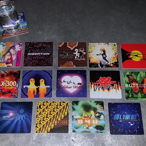 May include: A collection of 16 colorful square coasters with various designs. The coasters feature images of video game characters, logos, and abstract patterns. Some of the coasters have text on them, including "MAX 300", "Silent Hill", "Japan", "Butterfly", and "The Legend of Max 300".