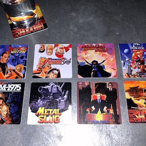 May include: Eight square coasters featuring retro video game artwork. The coasters feature images from the games: Art of Fighting, Samurai Shodown,  Street Fighter IV,  Metal Slug,  Real Bout Fatal Fury,  NAM-1975,  The  Hunter, and Magician Lord.