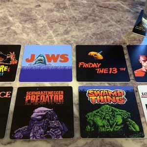 May include: Nine square coasters featuring retro video game graphics of popular horror movies. The coasters feature images of Freddy Krueger, Jaws, Jason Voorhees, the Terminator, Beetlejuice, Predator, Swamp Thing, and the Ghostbusters. The coasters are black with white and red text.