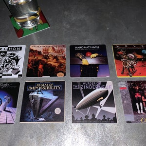 May include: Nine square coasters with album covers printed on them. The coasters feature a variety of album covers, including "Archon", "The Seventh Seal", "Hard Hat Mack", "Mule", "Realm of Impossibility", "Murder on the Zinderneuf", and "One on One".