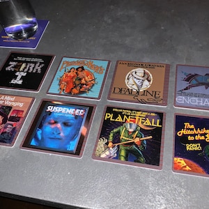 May include: Nine square coasters with different movie titles and images. The coasters are brown with a red border. The titles include "Zonk", "Planetfall", "Deadline", "Enchanter", "The Hitchhiker's Guide to the Galaxy", "A Mind Forever Voyaging", "Suspended", "Phantom Knights", and "A Locked Door A Dead Man".