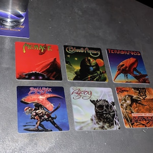 May include: Six square coasters with colorful sci-fi illustrations. The coasters feature images of spaceships, monsters, and futuristic landscapes. The titles of the coasters are "Menace", "Blood Money", "Terrorpods", "Ballrix", "Agony", and "Shadow of the Beast".