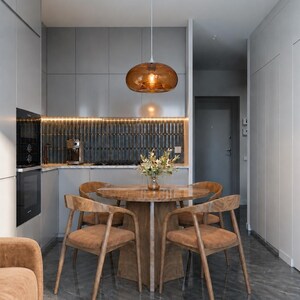 May include: A modern dining area with a round wooden table and four chairs. A brown glass pendant light hangs above the table. The room has gray cabinets and a dark gray tiled backsplash. The floor is a dark gray marble.
