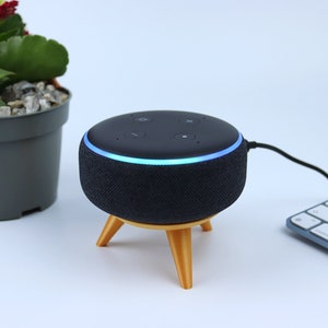 Peut inclure: Un haut-parleur intelligent noir avec un anneau lumineux bleu, posé sur un support en bois à trois pieds. Le haut-parleur est connecté à un clavier blanc.