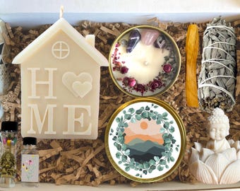 May include: A gift box filled with self-care items. Includes a house-shaped candle with "HOME" text, a candle with crystals and flowers, a coaster with a mountain scene, a sage bundle, a palo santo stick, a Buddha statue, and small bottles.