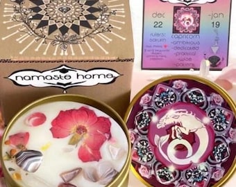 May include: A gold-colored candle tin with a white wax interior, adorned with a large red flower and gemstones. The candle is part of a set that includes a box with a geometric design and a card with the text "namaste home".