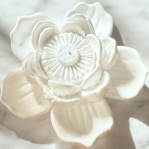 May include: A white ceramic flower sculpture with detailed petals and a textured centre. The decorative flower is likely for home decor, set against a marble-effect background. The sculpture is a detailed floral design.
