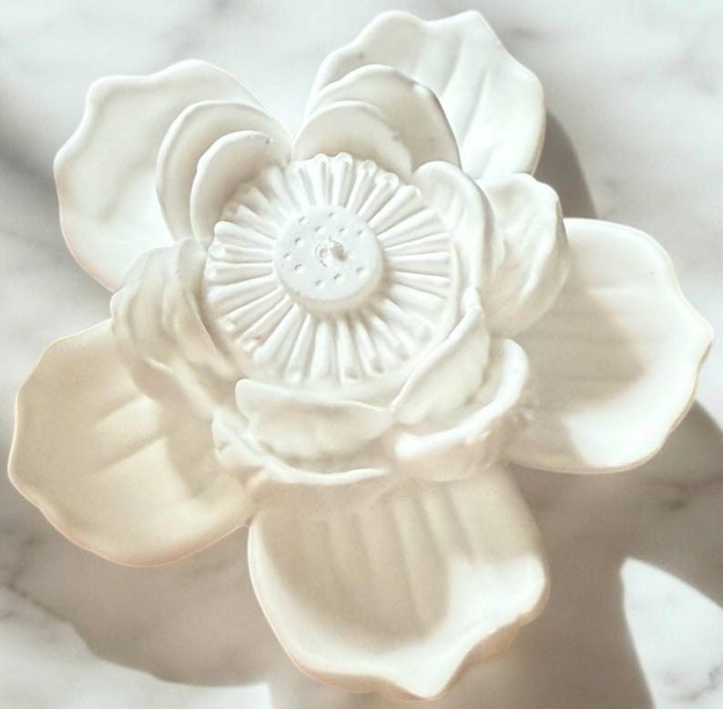 May include: A white ceramic flower sculpture with detailed petals and a textured centre. The decorative flower is likely for home decor, set against a marble-effect background. The sculpture is a detailed floral design.