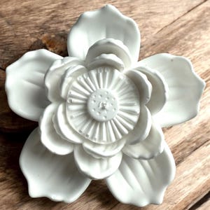 May include: A white, ceramic flower with multiple petals and a detailed center. The flower is set against a wooden background, creating a contrast in textures. The design is a close-up shot, highlighting the intricate details of the floral sculpture.