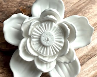 Puede incluir: Una escultura floral de cerámica blanca con pétalos detallados y un centro texturizado. La flor se encuentra sobre un fondo de madera. La toma de primer plano resalta los intrincados detalles del diseño floral.