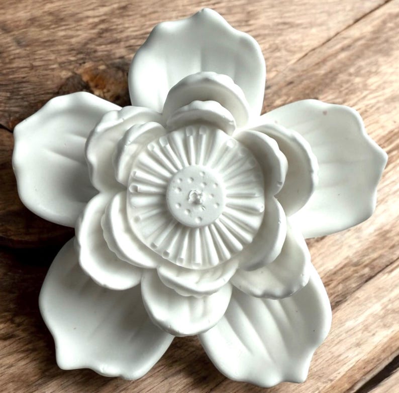 May include: A white ceramic flower sculpture with detailed petals and a textured centre. The flower is set against a wooden background. The close-up shot highlights the intricate details of the floral design, showcasing the craftsmanship of the piece.