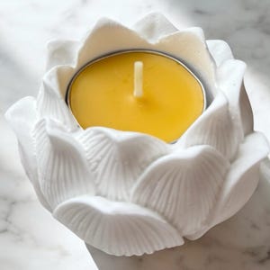 Puede incluir: Un portavelas de cerámica blanca con forma de flor de loto con una vela de té amarilla en su interior. El portavelas tiene diseños detallados de pétalos y una copa de metal. La vela tiene una mecha blanca.