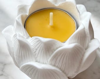 Puede incluir: Un portavelas de cerámica blanca con forma de flor de loto con una vela de té amarilla en su interior. El portavelas tiene diseños detallados de pétalos y una copa de metal. La vela tiene una mecha blanca.