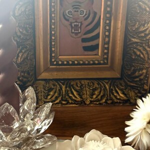 May include: A framed picture of a tiger with a fierce expression, set in an ornate gold frame. Decorative crystal and ceramic lotus flowers are in the foreground, with a white flower on the right.