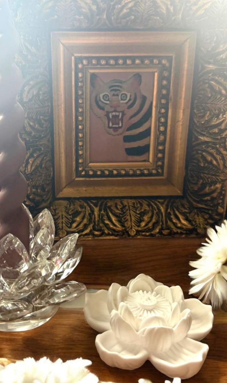 May include: A framed picture of a tiger with a fierce expression, set in an ornate gold frame. Decorative crystal and ceramic lotus flowers are in the foreground, with a white flower on the right.