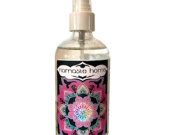 May include: Clear glass spray bottle with a white top and a label featuring the text "namaste home". The label has a black border and a colorful mandala design with pink, purple, and teal accents.