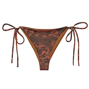 May include: A pair of bikini bottoms with a rust-colored background and a dark gray phoenix and floral pattern. The bottoms have tie closures on each side. The interior is a solid tan color.