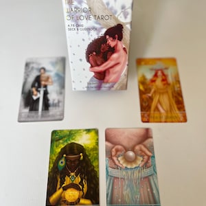 Puede incluir: Una baraja de cartas de tarot titulada "The Warrior of Love Tarot" con una caja que presenta una ilustraci&oacute;n de dos figuras abraz&aacute;ndose. Se muestran varias cartas de tarot, incluyendo "Ace of Pentacles" y "Ace of Cups". La baraja incluye 78 cartas y una gu&iacute;a.