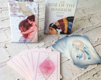 Puede incluir: Dos barajas de tarot, una titulada "The Warrior of Love Tarot" con una pareja abrazándose en la portada, y la otra titulada "The Rise of the Warrior Oracle" con una mujer con un tocado en la portada.