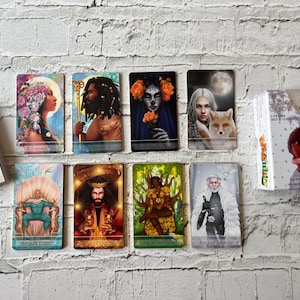 Puede incluir: Un mazo de cartas del tarot y varias cartas individuales se muestran sobre un fondo de ladrillos blancos. Las cartas presentan ilustraciones coloridas de figuras, incluyendo una emperatriz, un emperador y un rey. La caja del mazo muestra dos figuras abraz&aacute;ndose.
