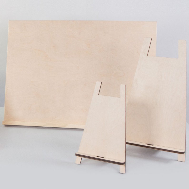 May include: A set of unfinished wooden easels and a large wooden panel. The easels are in different sizes, featuring a simple A-frame design. The wood has a light, natural colour, and the pieces are arranged against a plain background.
