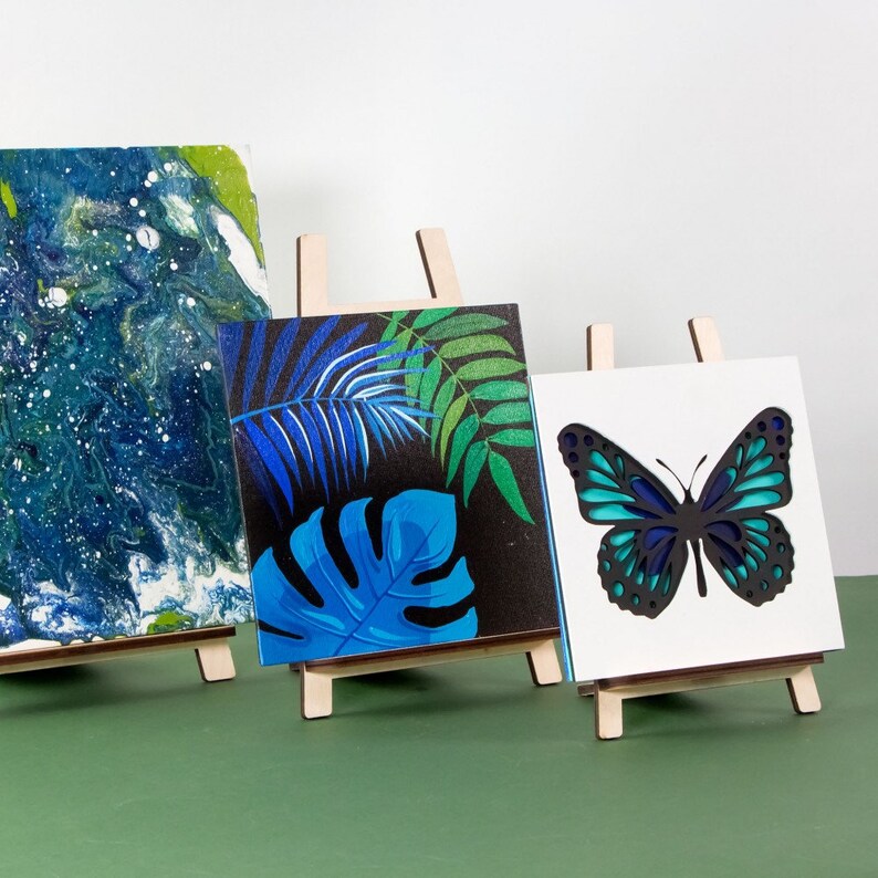May include: Three small artworks displayed on wooden easels. One has a blue and white abstract design, another shows blue and green tropical leaves on a black background, and the third features a layered butterfly design in blue and black.