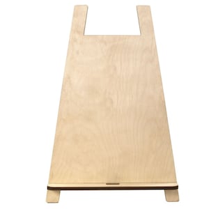 May include: A light-coloured wooden easel with a rectangular base and a wide, angled body. The top features a cutout handle. The wood grain is visible, and the easel is designed to hold items for display.