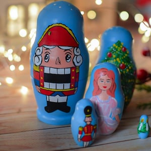 May include: A collection of hand-painted blue Matryoshka dolls. The largest doll features a Nutcracker design, while others depict a Christmas tree, a woman, and a soldier. The dolls are arranged on a wooden surface with blurred background lights.
