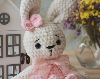 May include: A white crocheted bunny wearing a pink dress with a lace trim and a pink bow. The bunny has a pink flower on its head and blue eyes.