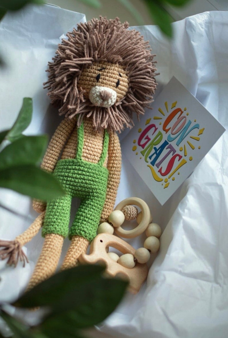 May include: A handmade crochet lion toy with a brown mane and green overalls. A wooden teether and a card that says "CONGRATS" are also in the box. The lion is a soft toy for babies.